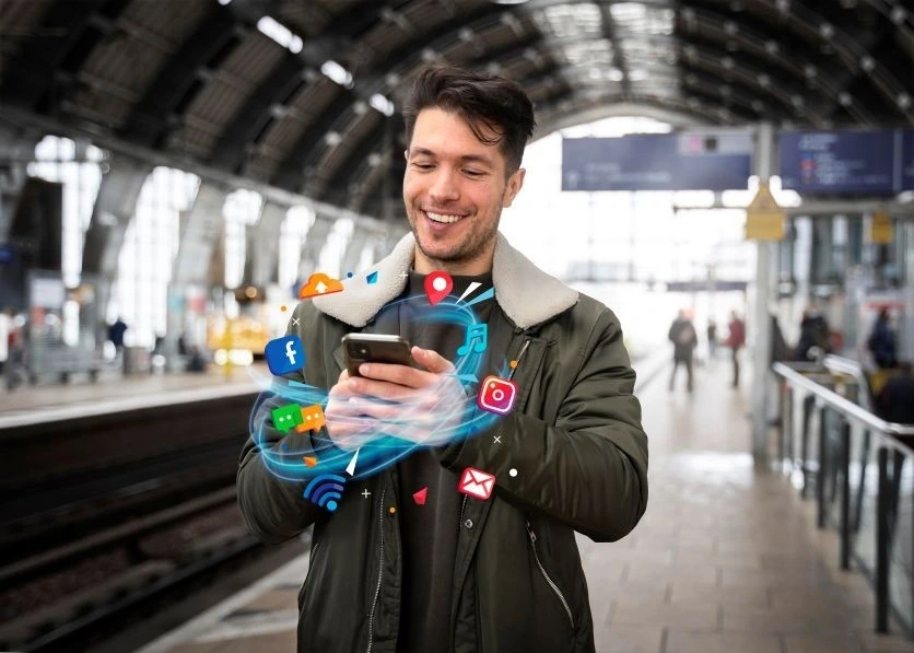 Top Digital Marketing Trends in 2025, smiling man at train station using smartphone surrounded by floating social media and online communication icons.