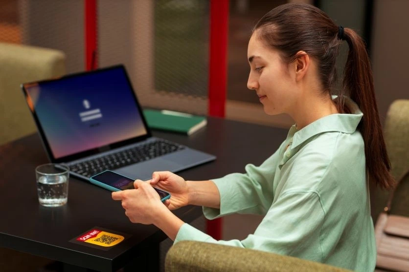 Young woman using smartphone at a desk with laptop in background, representing ways to earn money online with AI in 2025.